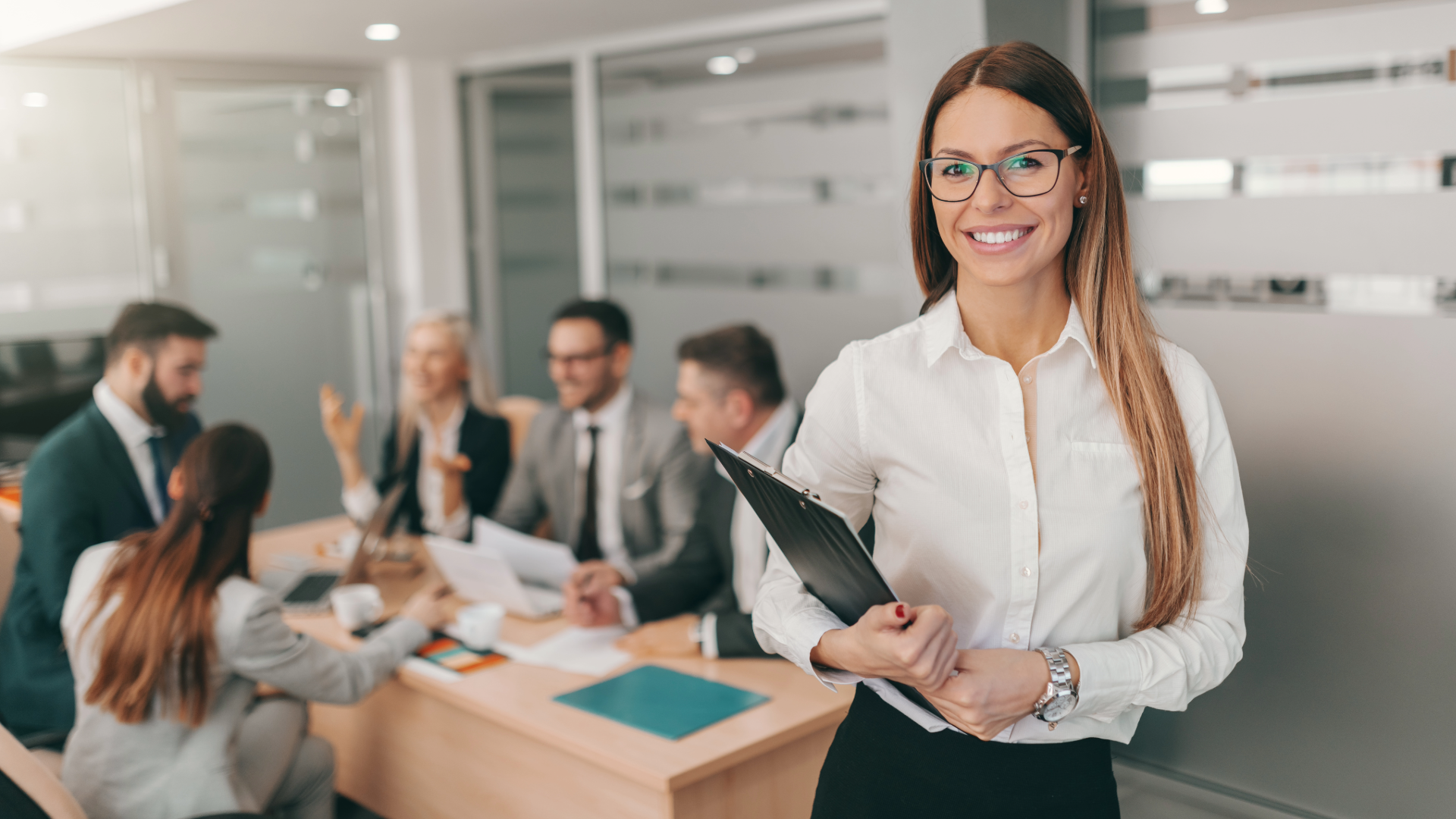 picture of a woman wearing glasses and a white dress shirt holding a clipboard with members of a team blurred our behind herew